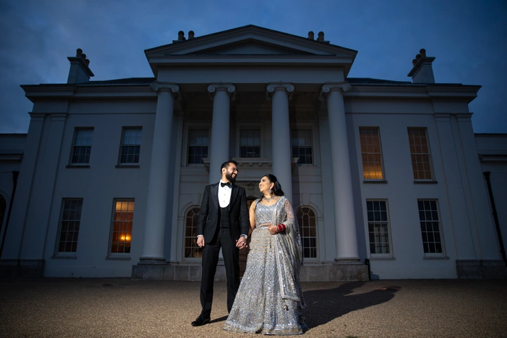 A bride and groom holding hands outside Hylands House in the evening. 