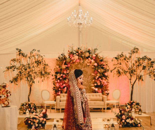 A bride in a flowing red dress posing in front of her red and pink floral decorations. 
