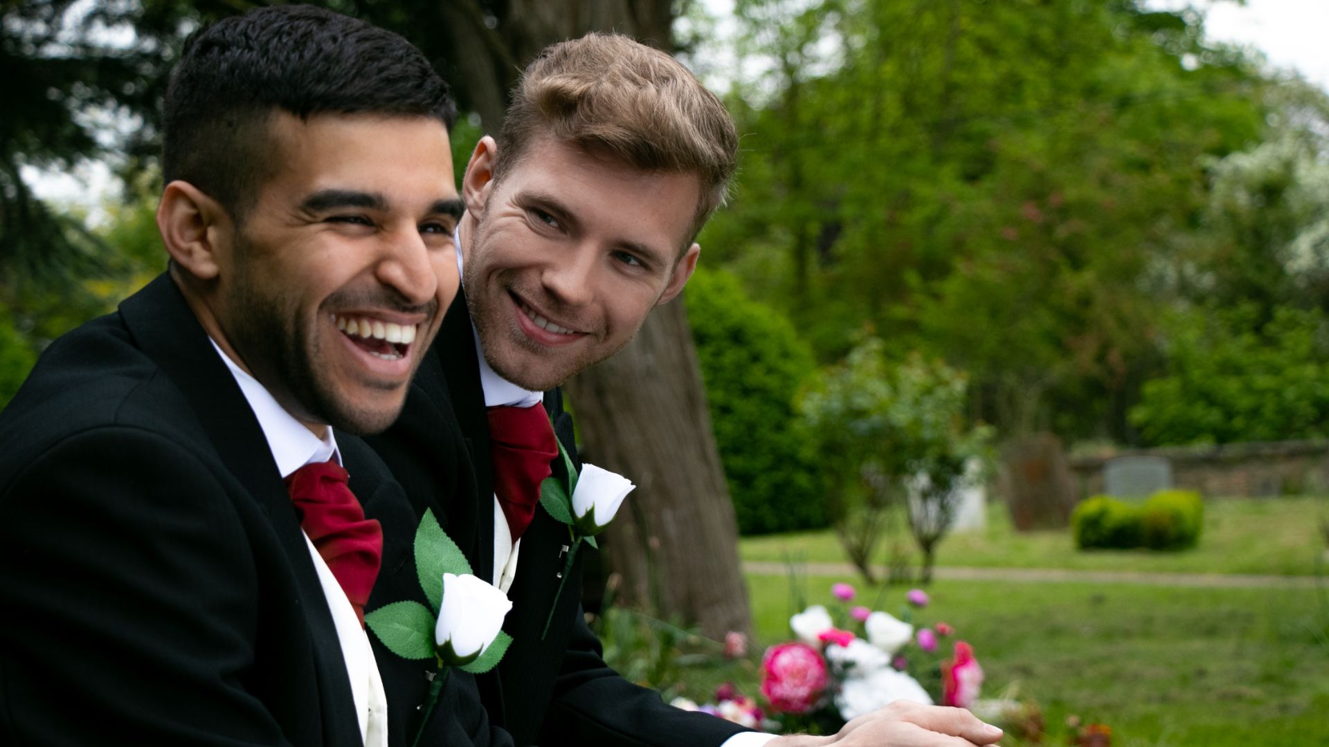 Two men wearing boutonnieres sitting next to each other outside and smiling. 