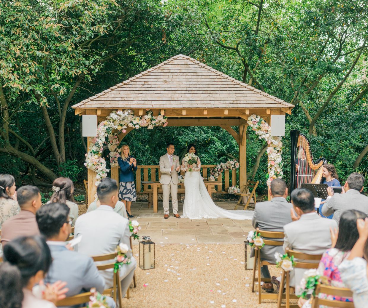 A couple getting married under the gazebo of the Outdoor Ceremony Garden which is adorned with light pink and white flowers. 