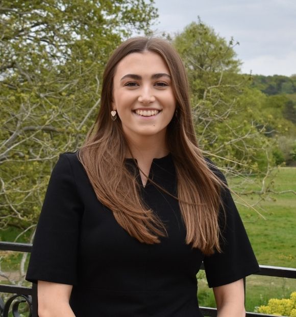 Grace, a woman with long brunette hair, smiling while standing on the East Terrace. 
