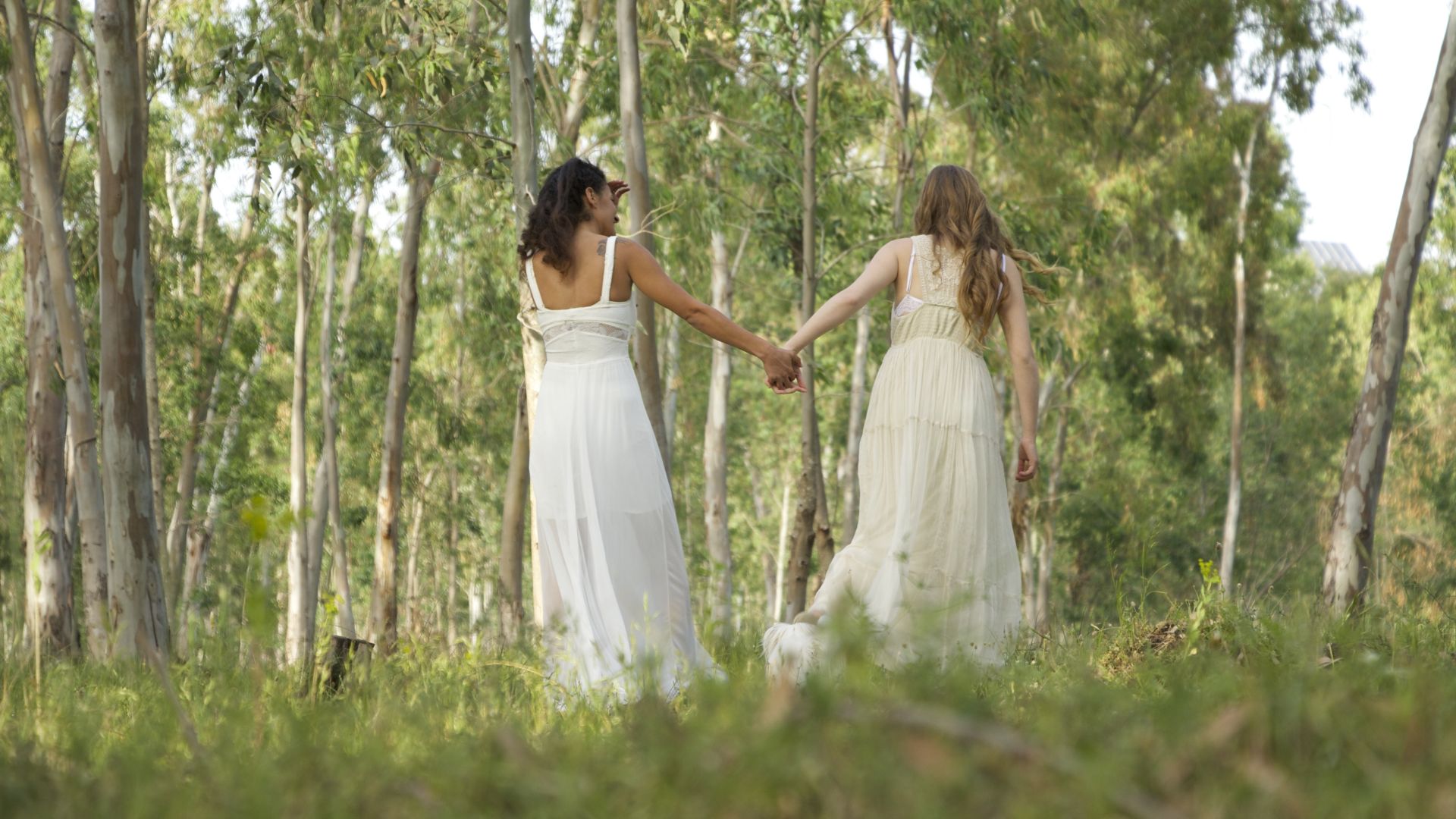 Two women wearing long white dresses and holding hands while walking outdoors. 