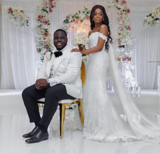 A groom sitting on a chair while his bride stands behind him with her hand on his shoulder. 