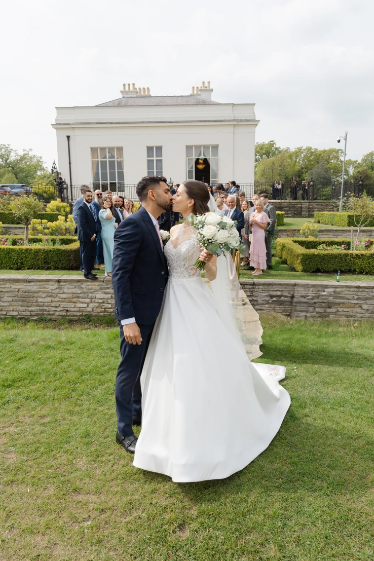 A bride and groom kissing outside Hylands House as their family and friends look on. 
