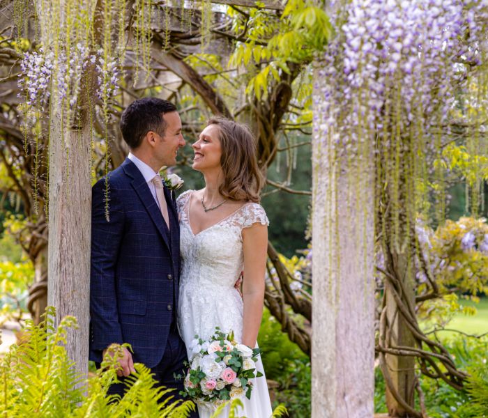 A bride and groom standing under the wisteria of the Pleasure Gardens.